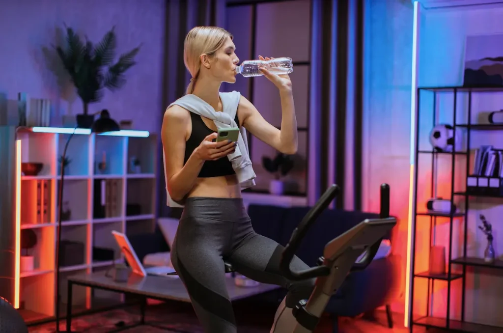 Woman drinking water in a gym and holding a mobile phone